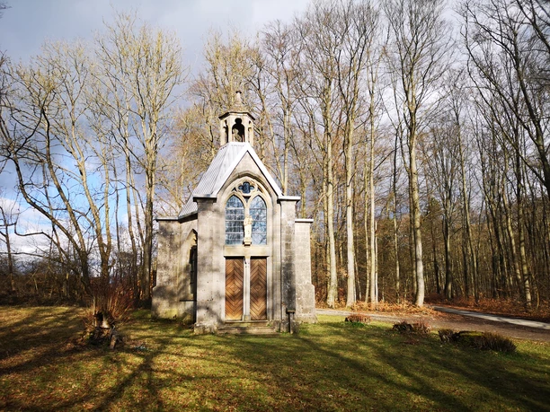 Kleine Steinkapelle mit Glockenturm, umgeben von Bäumen, steht im lichten Schatten eines Waldes.