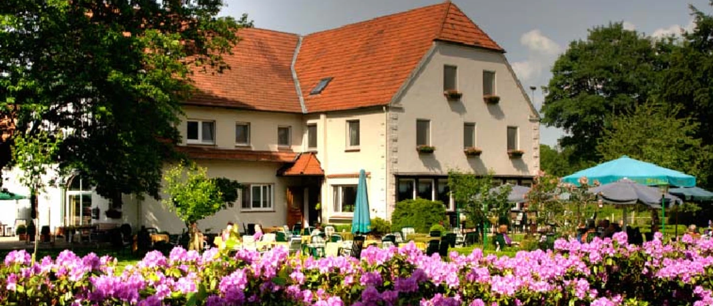 Ein großes Haus mit rotem Satteldach, umgeben von blühenden Rhododendren unter freiem Himmel.
