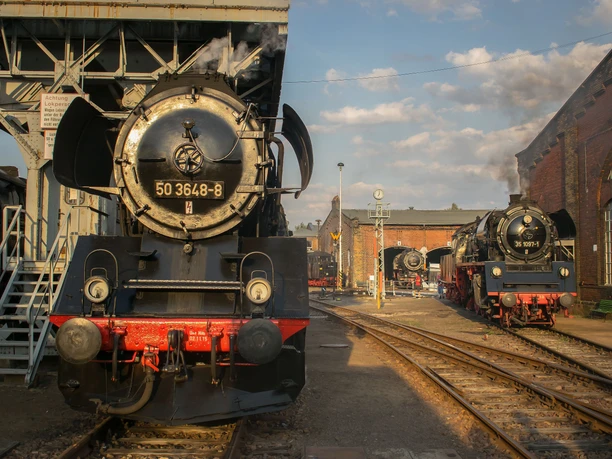 Heizhausfest_1_Johnny_Ullmann.jpg Historische Dampflokomotiven im Bahnbetriebswerk bei Sonnenuntergang, mit Personen links.