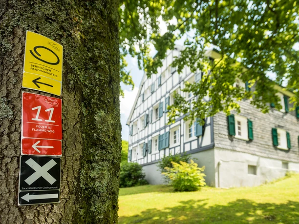 Wiedenest Ein Fachwerkhaus im Grünen, mit Wanderwegweisern an einem Baum im Vordergrund.