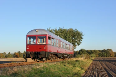 Bleckeder-kleinbahn-triebwagen-foto_ccbysa_foto©Bleckeder-kleinbahn.jpg Ein roter historischer Triebwagen der Bleckeder Kleinbahn fährt auf Gleisen durch eine ländliche Herbstlandschaft.