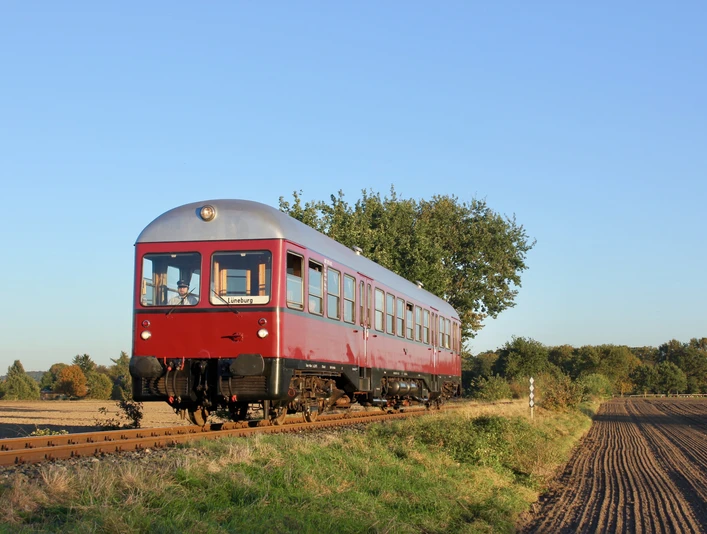 Bleckeder-kleinbahn-triebwagen-foto_ccbysa_foto©Bleckeder-kleinbahn.jpg Ein roter historischer Triebwagen der Bleckeder Kleinbahn fährt auf Gleisen durch eine ländliche Herbstlandschaft.