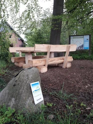 Holztisch und Bänke auf Waldboden nahe einem Wegweiser in einer ländlichen, grünen Umgebung.