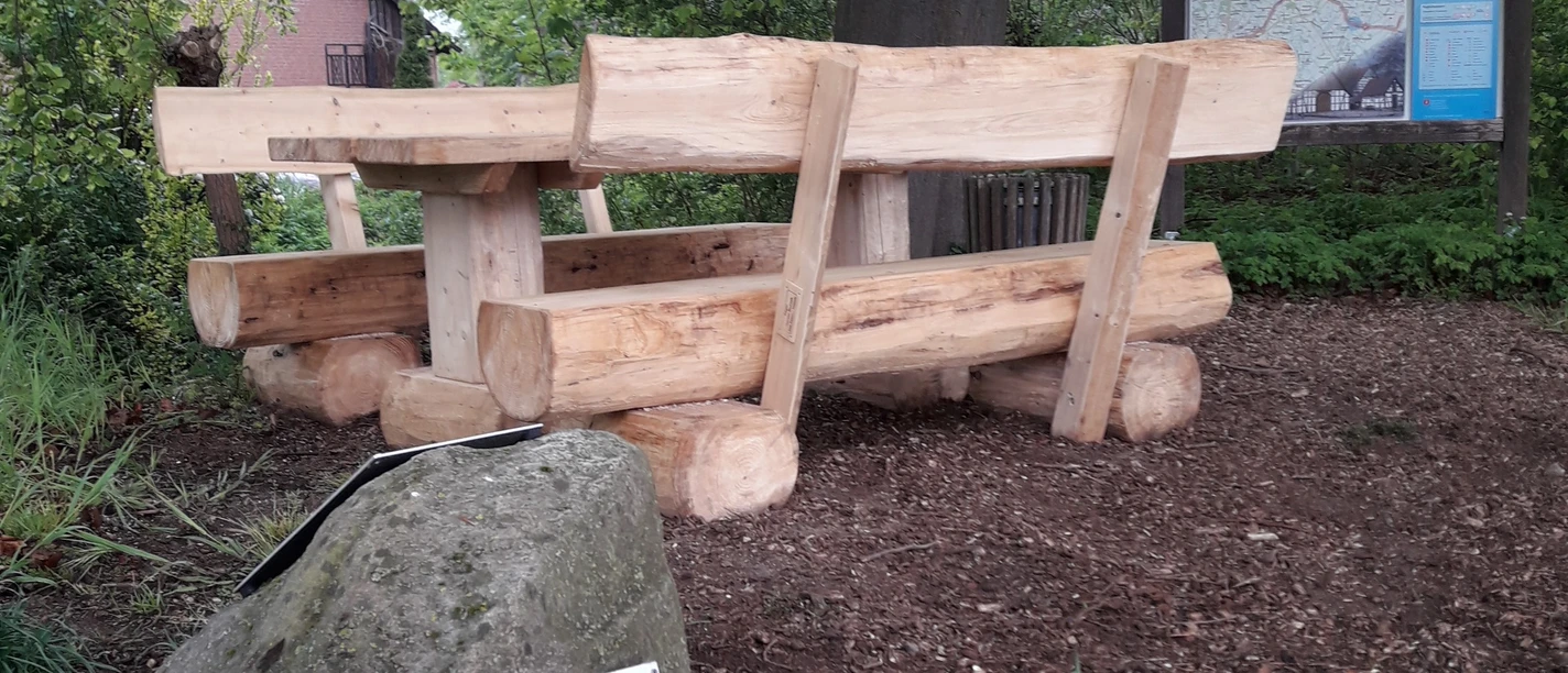 Holztisch und Bänke auf Waldboden nahe einem Wegweiser in einer ländlichen, grünen Umgebung.