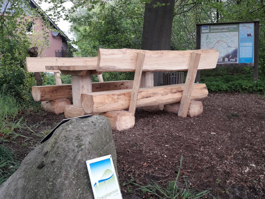 Sitzgruppe neu.jpg Holztisch und Bänke auf Waldboden nahe einem Wegweiser in einer ländlichen, grünen Umgebung.