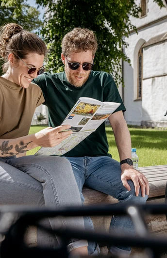 Grenzgaengerroute_Versmold Radkarte_Christoph Steinweg_1_teuto.jpg Ein lächelndes Paar sitzt auf einer Bank im Grünen, betrachtet eine Karte, Fahrräder daneben.