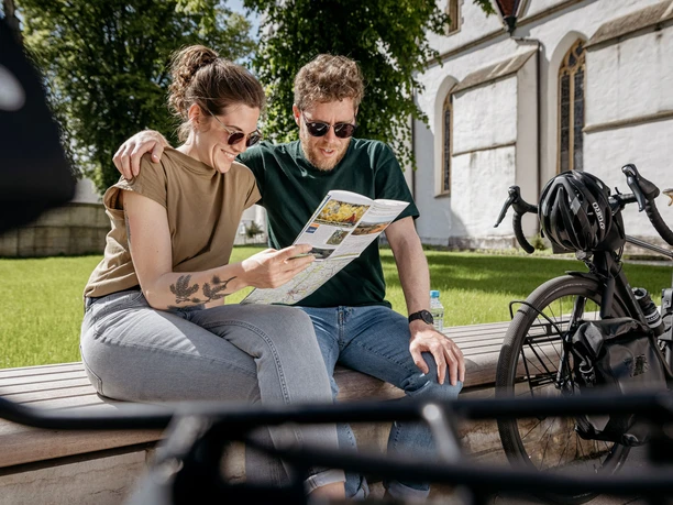 Grenzgaengerroute_Versmold Radkarte_Christoph Steinweg_1_teuto.jpg Ein lächelndes Paar sitzt auf einer Bank im Grünen, betrachtet eine Karte, Fahrräder daneben.