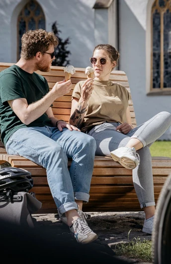 Grenzgaengerroute_Versmold_Christoph Steinweg_3_teuto.jpg Ein Paar sitzt auf einer Holzbank im Park und genießt Eis, mit Fahrrädern im Vordergrund.