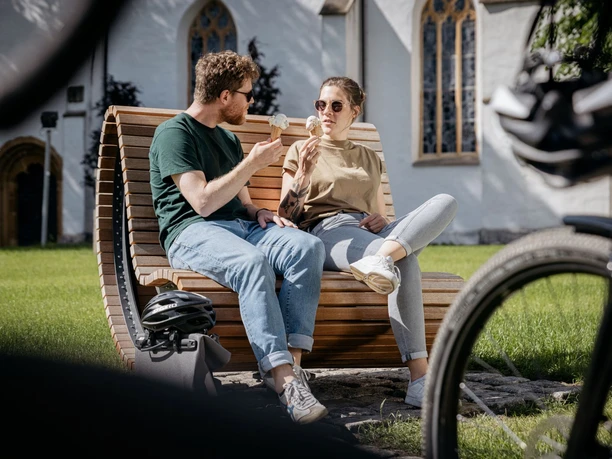 Grenzgaengerroute_Versmold_Christoph Steinweg_3_teuto.jpg Ein Paar sitzt auf einer Holzbank im Park und genießt Eis, mit Fahrrädern im Vordergrund.