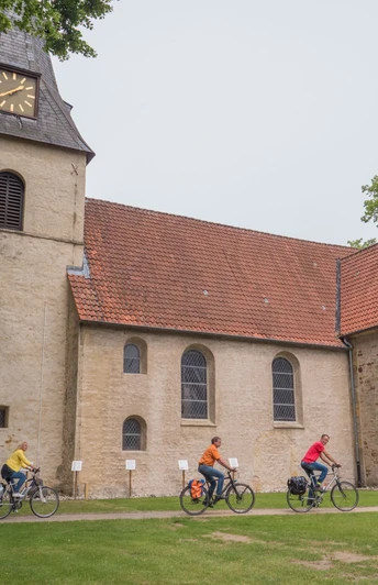 84_Grenzgängerroute_Bockhorst_Max Daerr_2018_teuto.jpg Eine Gruppe von Radfahrern fährt an einer historischen Kirche mit rotem Ziegeldach vorbei.
