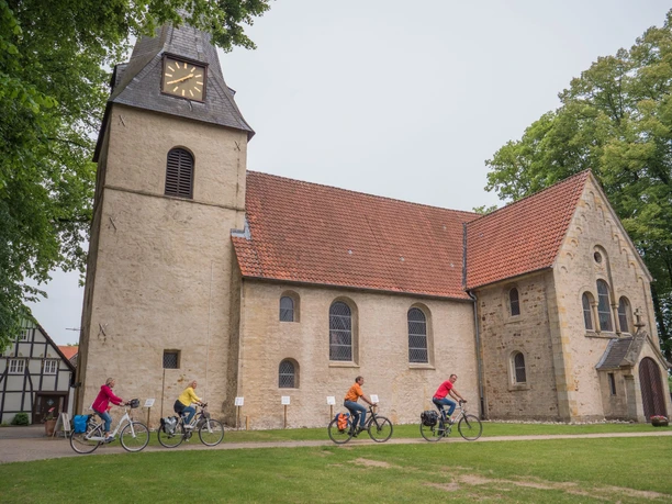84_Grenzgängerroute_Bockhorst_Max Daerr_2018_teuto.jpg Eine Gruppe von Radfahrern fährt an einer historischen Kirche mit rotem Ziegeldach vorbei.