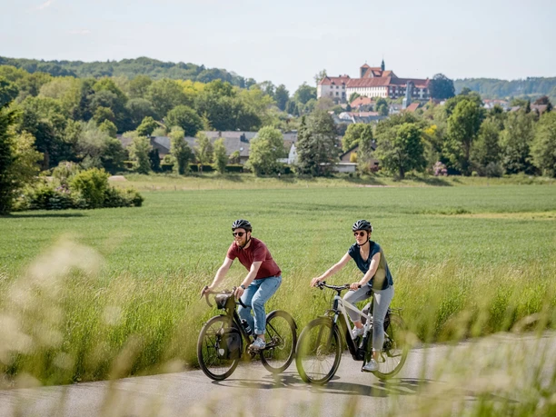 Zwei Radfahrer genießen eine ländliche, grüne Landschaft mit einem Schloss im Hintergrund bei sonnigem Wetter.