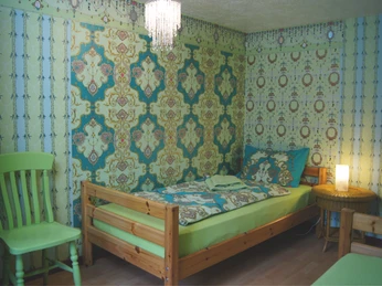 Eichenhof-Kinderzimmer-App-2 Ein Kinderzimmer mit Holzbett, farbenfroher Tapete, grünem Stuhl und Deckenlampe.