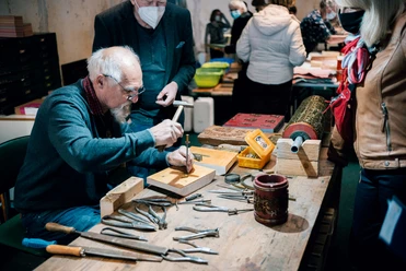 Druckerbande aus Einbeck Ein Mann prägt mit Hammer und Werkzeug Metallplatten, umgeben von Zuschauern in einer Werkstatt.