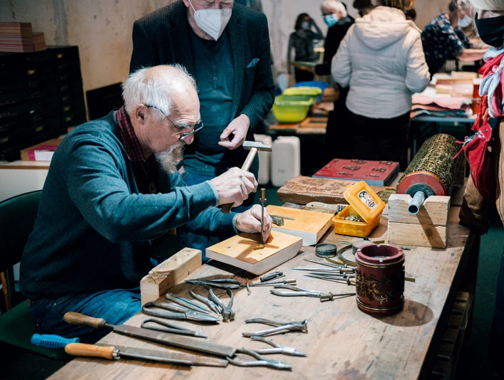 Druckerbande aus Einbeck Ein Mann prägt mit Hammer und Werkzeug Metallplatten, umgeben von Zuschauern in einer Werkstatt.