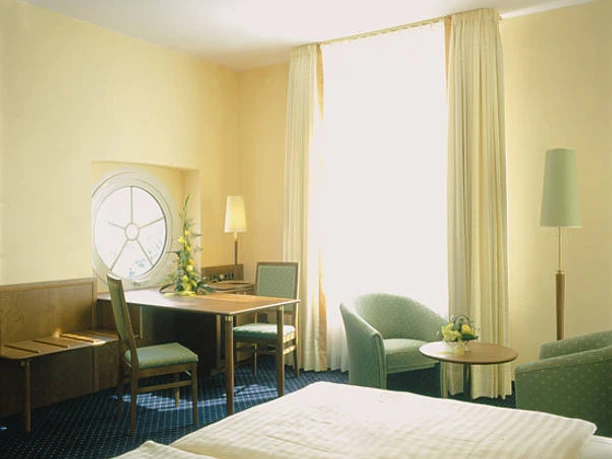 Doppelzimmer-Deluxe 01.jpg Helles Doppelzimmer mit zwei Sesseln, Tisch und einem großen Fenster, das viel Licht hereinlässt.