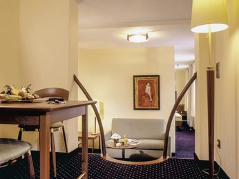 JuniorSuite 01.jpg Junior Suite mit elegantem Holzstuhl, Couchtisch und Sofa, Kunstwerk an der Wand, warmes Licht.