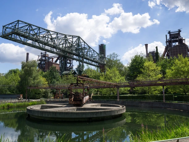 Landschaftspark Duisburg-Nord Klärteich und Krokodil, Duisburg Im Landschaftspark Duisburg-Nord zeigt das Bild den Klärteich mit einer zentral gelegenen Metallbrücke, umgeben von industriellen Strukturen und üppiger Vegetation.
