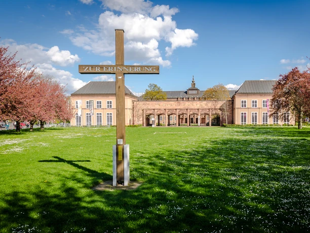 View of the wooden cross that commemorates the destroyed St. John's Church and the building of the Grassi Museums of Ethnology Musical Instruments and Applied Arts on Johannisplatz, where you can see wonderful exhibitions, cultural institution in Leipzig, museum, history, culture