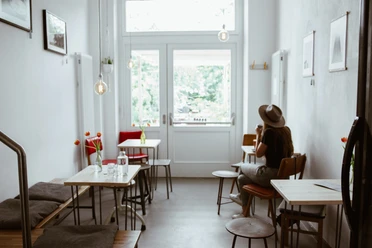 Innenansicht Café Analog - Cafés in Leipzig Zusehen sind die Sitzplätze des Cafés: Kleine Tische und Bänke aus Holz sowie kleine Bilder, die an der Wand hängen. Der Raum wird von einem großen Glasfenster erhellt, Cafe, Gastronomie, Freizeit