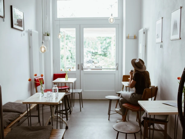 Innenansicht Café Analog - Cafés in Leipzig Zusehen sind die Sitzplätze des Cafés: Kleine Tische und Bänke aus Holz sowie kleine Bilder, die an der Wand hängen. Der Raum wird von einem großen Glasfenster erhellt, Cafe, Gastronomie, Freizeit