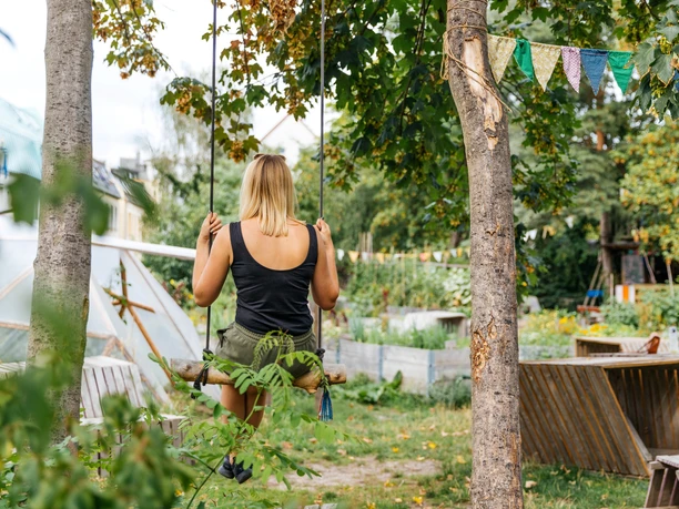 Besucherin im Gemeinschaftsgarten Annalinde - Urbane Kultur in Leipzig Blick auf die vielseitige Gartenidylle des Annalinde, auf einer Schaukel zwischen zwei Bäumen sitzt eine junge Frau, den Blick in den Garten gewandt, Annalinde, Urbane Landwirtschaft