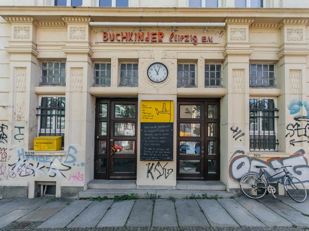 Pohled na fasádu a exteriér Buchkinder e.V. za denního světla, s vývěsním štítem, nákupy, kultura