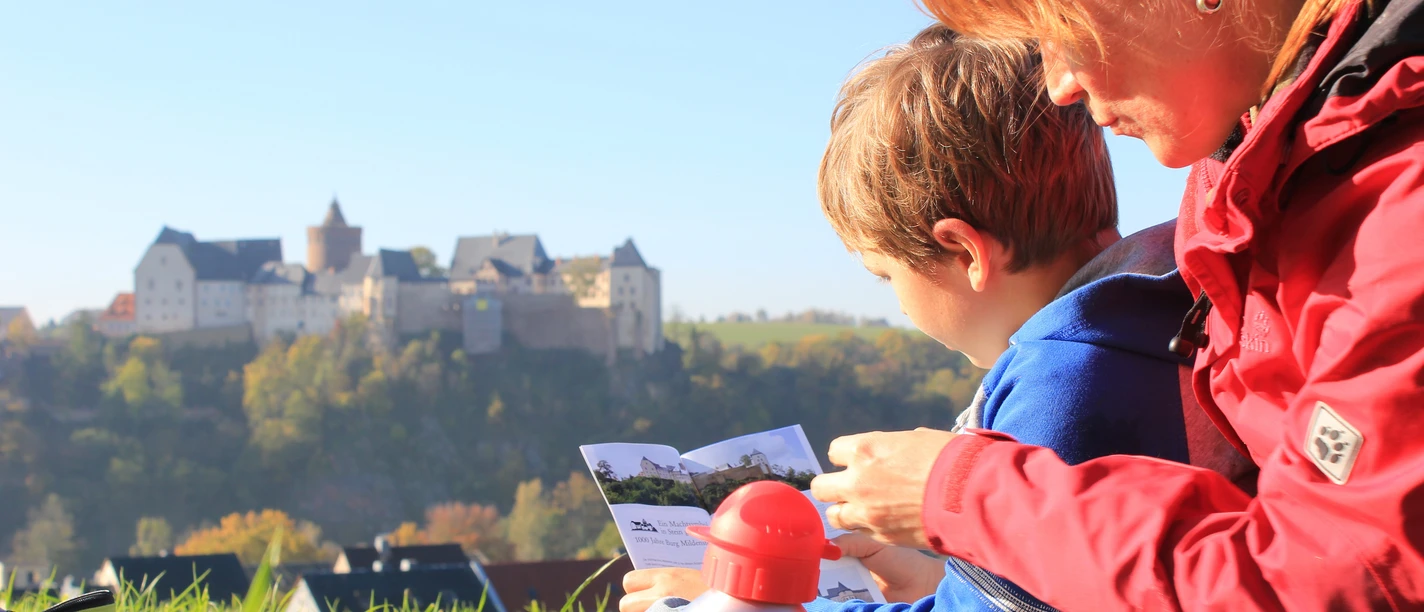 Burg Mildenstein - Sehenswürdigkeiten in der Leipzig Region Eine Frau mit Kind sitzt mit einer Broschüre in der Hand auf einer Wiese, im Hintergrund erhebt sich Burg Mildenstein, Auflug, Familie,
