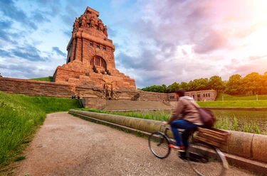 Völkerschlachtdenkmal - Sehenswürdigkeiten in Leipzig Blick auf das von der warmen Sonne angestrahlte Völkerschlachtdenkmal Leipzig, während ein Fahrradfahrer am See der Tränen vorbeiradelt, eine wahre Sehenswürdigkeit und ein besonderes Stück der Architektur in Leipzig, Ausflug, Sehenswürdigkeiten, Kultur