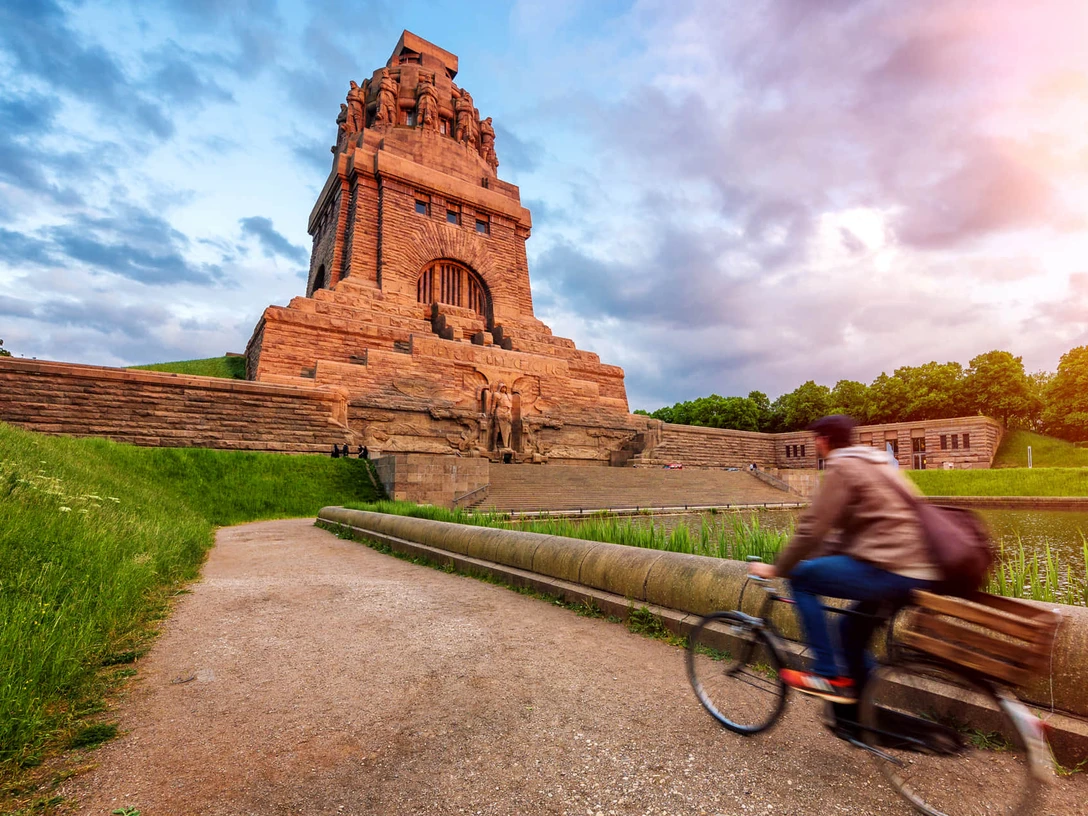 Völkerschlachtdenkmal - Sehenswürdigkeiten in Leipzig Blick auf das von der warmen Sonne angestrahlte Völkerschlachtdenkmal Leipzig, während ein Fahrradfahrer am See der Tränen vorbeiradelt, eine wahre Sehenswürdigkeit und ein besonderes Stück der Architektur in Leipzig, Ausflug, Sehenswürdigkeiten, Kultur