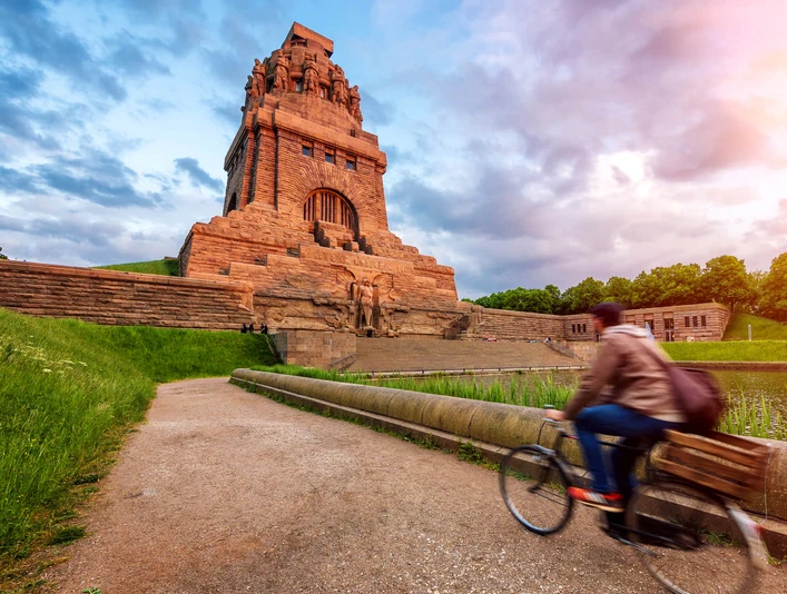 Völkerschlachtdenkmal - Sehenswürdigkeiten in Leipzig Blick auf das von der warmen Sonne angestrahlte Völkerschlachtdenkmal Leipzig, während ein Fahrradfahrer am See der Tränen vorbeiradelt, eine wahre Sehenswürdigkeit und ein besonderes Stück der Architektur in Leipzig, Ausflug, Sehenswürdigkeiten, Kultur