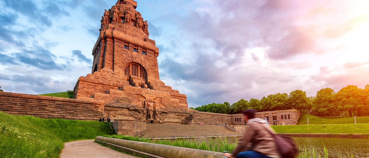 Pohled na Völkerschlachtdenkmal Leipzig, osvětlený hřejivým sluncem, zatímco cyklista projíždí kolem jezera slz, skutečné památky a zvláštního architektonického díla v Lipsku, Výlet, Památky, Kultura