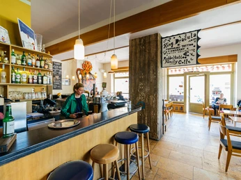 Café Cantona - Gastronomie Leipzig Gastraum des Café Cantona mit langer Bar und Barstühlen sowie einer großen Fensterfront mit Blick auf die Windmühlenstraße