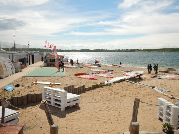 Sportovci, kteří si chtějí vyzkoušet windsurfing na jezeře, stojí na břehu jezera Schladitz.