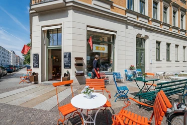 Vue sur la terrasse du glacier Pfeifer avec différents meubles de jardin et façade de style "Gründerzeit" (époque des fondateurs), espace libre pour les enfants.