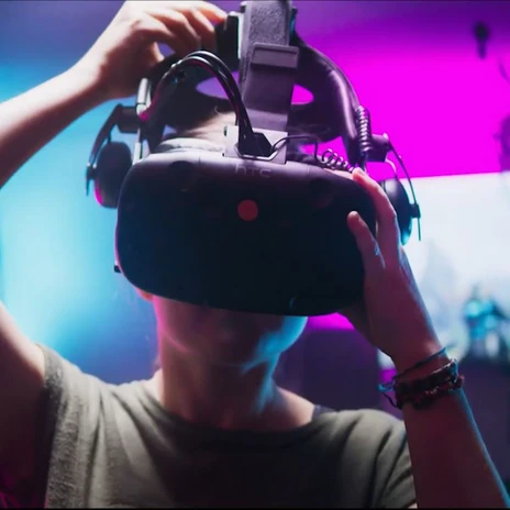 Eine Person mit VR-Brille in einem farbig beleuchtetem Escape-Room, Monitore im Hintergrund.