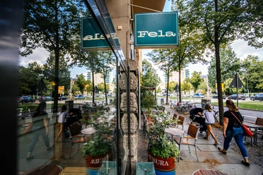 Widok wzdłuż fasady i dużej witryny restauracji Fela, na niebieski szyld sklepu, ulicę i odbicie w oknie; w letni dzień, miejsce do siedzenia na zewnątrz, restauracja, gastronomia