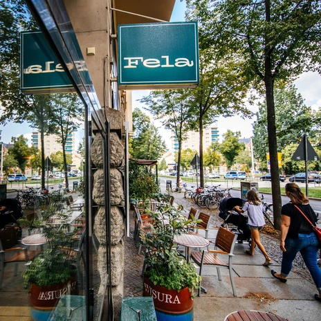 Widok wzdłuż fasady i dużej witryny restauracji Fela, na niebieski szyld sklepu, ulicę i odbicie w oknie; w letni dzień, miejsce do siedzenia na zewnątrz, restauracja, gastronomia