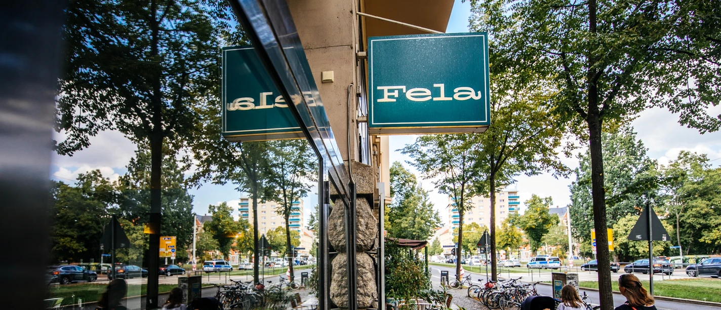Fela - Gastronomie in Leipzig Blick entlang der Fassade und des großen Schaufensters des Fela Restaurants, auf das blaue Geschäftsschild, den Straßenzug und die Spiegelung im Fester; an einem Sommertag, freisitz, restaurant, grastronomie