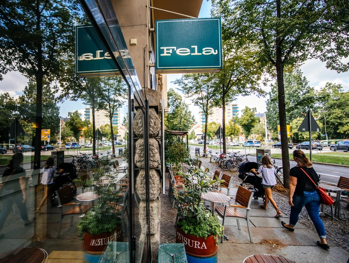 Fela - Gastronomie in Leipzig Blick entlang der Fassade und des großen Schaufensters des Fela Restaurants, auf das blaue Geschäftsschild, den Straßenzug und die Spiegelung im Fester; an einem Sommertag, freisitz, restaurant, grastronomie