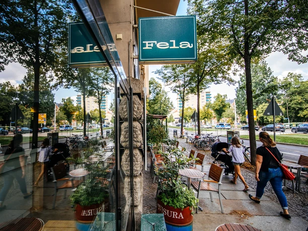 View along the façade and the large shop window of the Fela Restaurant, of the blue store sign, the street and the reflection in the window; on a summer's day, outdoor seating, restaurant, gastronomy