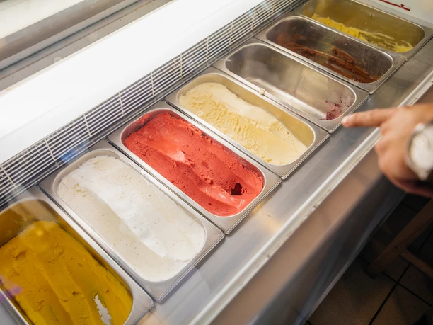 Gustav H. - Eisdielen & Eiscafés in Leipzig Blick in die bunte Eisvitrine und die darin gebotene Auswahl an Sorten; rechts ragt ein zeigender Finger eines Kunden in das Bild, eisdiele, eiscafe