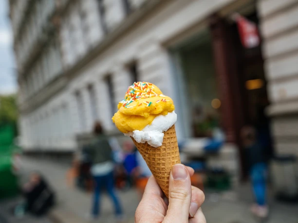 Gustav H. - Eisdielen & Eiscafés in Leipzig Fotografie zweier Kugeln Eis in einer Waffel, die von einer Hand gehalten werden; im Hintergrund sieht man den Eingang und die Fassade des Gustav H., eisdiele, eiscafe
