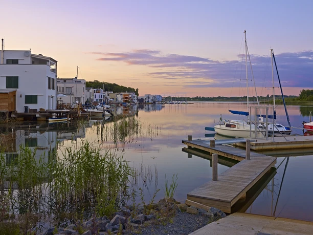 Un lac calme au coucher du soleil, bordé de péniches blanches modernes et de voiliers amarrés dans la baie.