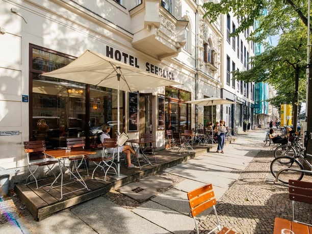 Blick auf den Außenbereich des Hotel Seeblicks, den Gehweg, die Häuserfassade und mehr mit Sonnenschirmen, Stühlen und Co. an einem Sommertag; mit Blick auf den Straßenzug und angrenzende Häuser, Gastronomie, kulinarik, restaurant, freisitz