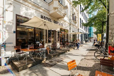 Hotel Seeblick - Gastronomie v Lipsku Pohled na venkovní areál hotelu Seeblick, chodník, fasádu domu a další místa se slunečníky, židlemi a podobně v letním dni; s výhledem na ulici a sousední domy, gastronomie, kulinářství, restaurace, venkovní posezení