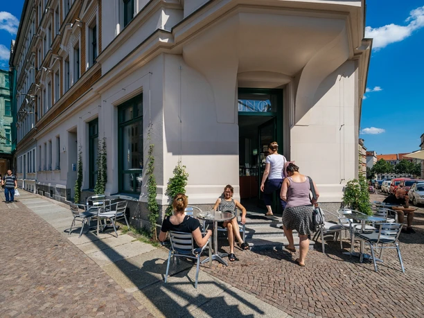 INO Café - Kavárny v Lipsku Pohled na vchod do kavárny v horní části rohové budovy a venkovní posezení před kavárnou s návštěvníky; na slunci, venkovní posezení, kavárna, gastronomie