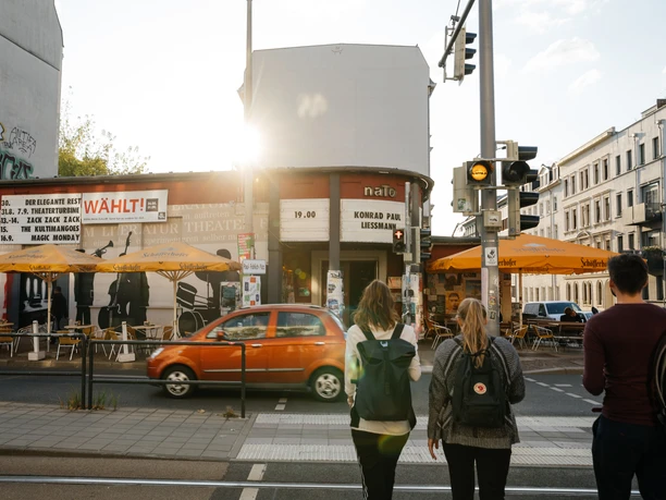 Przed centrum społeczno-kulturalnym naTo przy Karl-Liebknecht-Straße dwie kobiety i mężczyzna stoją na światłach na przejeździe kolejowym, aby przejść przez ulicę