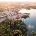 Blick von oben auf den Kiessee Eilenburg Die Luftaufnahme zeigt den Kiessee Eilenburg umgeben von üppigem Grün und einer Siedlung mit Wohnhäusern und Zelten unter einem sanften Tageslicht. Der See strahlt eine ruhige, einladende Atmosphäre aus.
