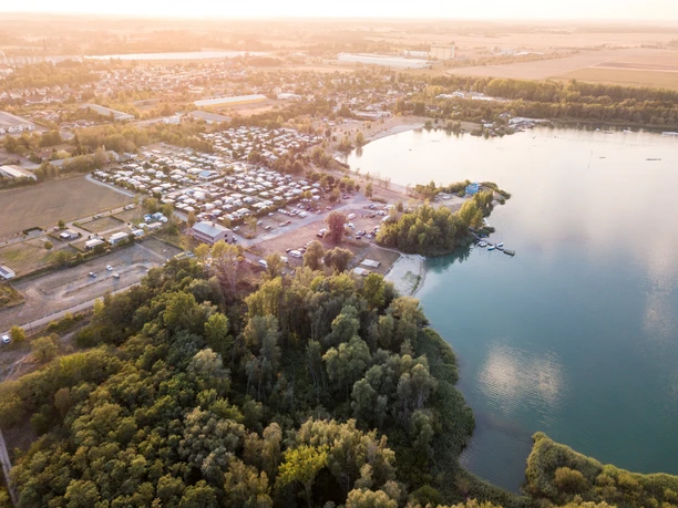 La vue aérienne montre le lac de gravier d'Eilenburg entouré d'une verdure luxuriante et d'un lotissement de maisons d'habitation et de tentes sous une douce lumière du jour. Le lac dégage une atmosphère calme et accueillante.